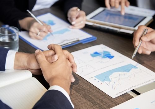 Crop shot of coworking businesspeople sitting at table with paper charts and tablet having meeting